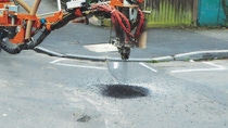 A road repair machine with a large, articulated arm is patching a pothole on an asphalt street. Nearby are houses and a fenced yard.