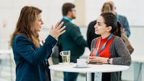 Two women engage in a lively discussion at a conference, standing by a table with drinks. Background shows other attendees.