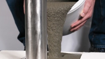 A person pours concrete from a white bucket into a mold. The smooth, metallic structure stands left, with jeans visible in the background. A person pours concrete from a white bucket into a mold. The smooth, metallic structure stands left, with jeans visible in the background.