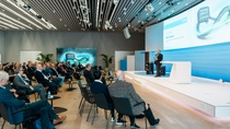 A man is speaking at a podium on a stage in a modern conference room (D105, Ludwigshafen). Attendees sit facing him, engaged.