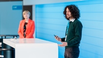 A man with curly hair speaks confidently at a podium, holding a microphone. A woman in an orange blazer listens attentively.