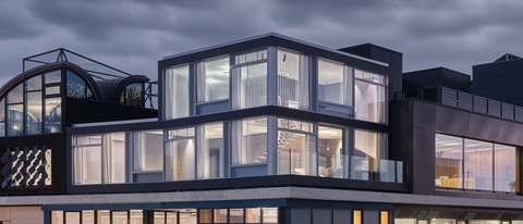 Modern building with glass walls and geometric shapes under a cloudy sky. Structure blends curved and angular elements.
NEST innovation and research building in Dübendorf, Switzerland, featuring upcycling solutions. The image shows the STEP2 unit with lights on, contrasting a cloudy, dark sky. Credit: Zooey Braun