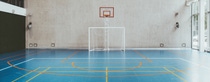 Spacious indoor sports hall with blue flooring, orange and yellow court lines. A soccer goal beneath a basketball hoop against the tall, beige wall. Bright, calm atmosphere. Spacious indoor sports hall with blue flooring, orange and yellow court lines. A soccer goal beneath a basketball hoop against the tall, beige wall. Bright, calm atmosphere.