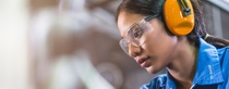 A woman in a blue uniform, safety goggles, and orange earmuffs focuses intently on industrial work.