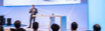Blurry image of a speaker on stage in a conference room. An audience is seated in front, with a modern podium and presentation screen in the background.