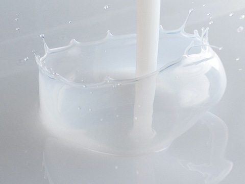 A stream of milky liquid splashes into a smooth, white surface, forming a circular crown with droplets suspended mid-air.