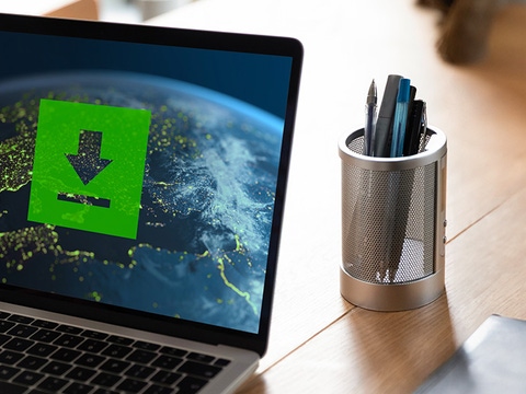 Woman using a laptop displaying a world map with a green download icon overlay. Bright workspace with pens in a holder, evoking a tech-focused atmosphere.