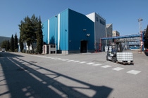 Industrial scene with a blue BASF building, a forklift carrying a large container, and shadowed walkways. Tall trees and clear sky in the background. Industrial scene with a blue BASF building, a forklift carrying a large container, and shadowed walkways. Tall trees and clear sky in the background.