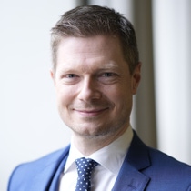 A man in a blue suit and polka dot tie smiles confidently indoors. The background is softly blurred.