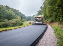 Construction workers in orange attire operate a paving machine, laying fresh asphalt on a rural road bordered by lush greenery and overcast skies. Construction workers in orange attire operate a paving machine, laying fresh asphalt on a rural road bordered by lush greenery and overcast skies.