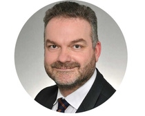 Smiling bearded man in a suit and tie, looking directly at the camera. The background is a simple gradient.