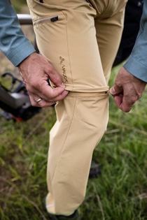 A person outdoors unzips the lower part of beige hiking pants. Green grass and equipment in the background suggest an adventure. A person outdoors unzips the lower part of beige hiking pants. Green grass and equipment in the background suggest an adventure.