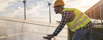 A man with a construction hat and reflective vest inspects solar panels at a solar farm during sunset. Wind turbines stand in the background under a partly cloudy sky.