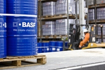Blue chemical barrels labeled "BASF" on a pallet in a warehouse. A worker in an orange uniform and helmet operates a forklift nearby.