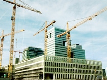 Four tall yellow cranes surround a modern building under construction against a blue sky.