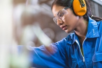 A woman in a blue uniform, safety goggles, and orange earmuffs focuses intently on industrial work.