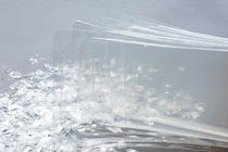 Clear translucent plastic sheets are stacked alongside scattered plastic flakes on a gray surface. Clear translucent plastic sheets are stacked alongside scattered plastic flakes on a gray surface.