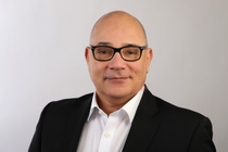 Bald man wearing glasses and a black suit with a white shirt, smiles subtly against a light gray background. Bald man wearing glasses and a black suit with a white shirt, smiles subtly against a light gray background.