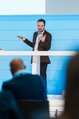 Man in a dark suit gives a presentation in front of a blue wall. He gestures with one hand while holding a clicker in the other.